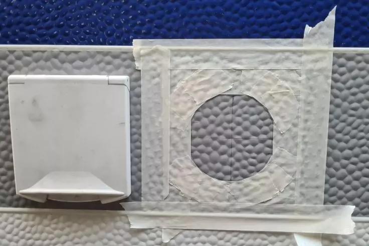 Graue, genoppte Wandfl&auml;che mit links einer wei&szlig;en Abdeckung mit Klappe und rechts einem runden Loch, das mit wei&szlig;en Klebebandstreifen umrahmt ist