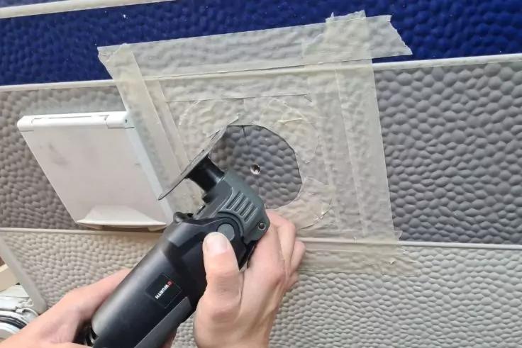 H&auml;nde halten ein elektrisches Schneidewerkzeug und schneiden ein rundes Loch in eine graue Wandverkleidung; Klebeband umgibt den Bereich, links eine wei&szlig;e Steckdosenabdeckung