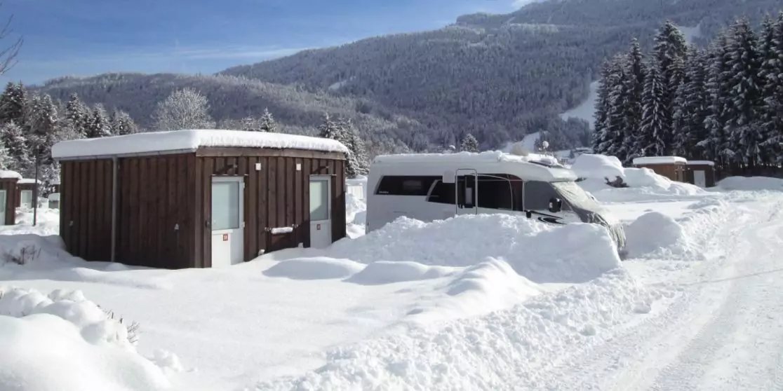 Kleine Holzh&uuml;tte und teilweise vom Schnee bedecktes Wohnmobil, gro&szlig;e Schneehaufen, verschneite Tannen und Berge unter blauem Himmel