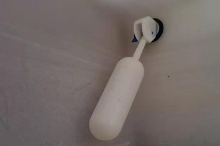 Wei&szlig;er l&auml;nglicher ovaler Kunststoffk&ouml;rper an einem wei&szlig;en Arm, befestigt mit einer runden schwarzen Saugnapfhalterung an einer hellen, gefliesten Wand