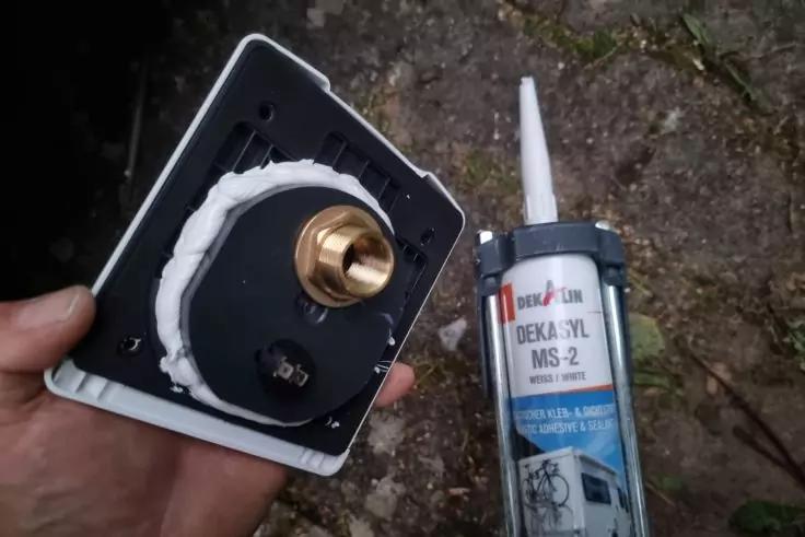 Hand h&auml;lt wei&szlig;e Abdeckplatte mit schwarzer R&uuml;ckseite und messingfarbener Einschraubmutter; wei&szlig;er Dichtstoff am Rand; daneben eine Dichtstoffkartusche auf steinigem Untergrund