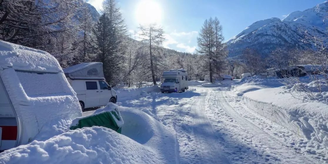 Schneebedeckter Campingplatz mit Wohnwagen, schneebedeckten B&auml;umen, Fahrspuren im Schnee, Bergen im Hintergrund und Sonne am Himmel