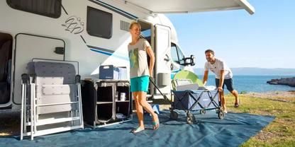 Wohnmobil mit ausgefahrener Markise am Meer, zwei Personen davor, Klappst&uuml;hle, tragbare Campingk&uuml;che und ein Wagen mit Ausr&uuml;stung auf einer blauen Bodenplane, Meer und Felsen im Hintergrund