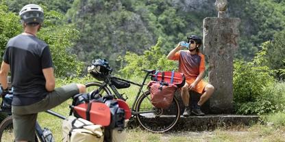 Zwei Radfahrende mit beladenen Reiser&auml;dern an einem Weg; eine Person sitzt auf einem steinernen Brunnen und trinkt aus einer Flasche, die andere steht neben dem Fahrrad. Bewaldete H&uuml;gel im Hintergrund
