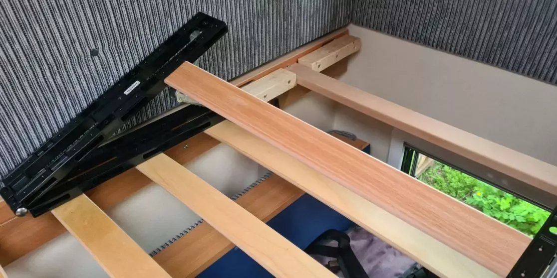 Holzlatten eines Bettgestells, eine lange Holzplanke quer darauf, schwarzer Metallbeschlag, geriffelte graue Wandverkleidung oben, seitliches Fenster mit gr&uuml;nen Pflanzen drau&szlig;en