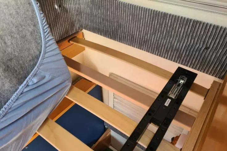 Halb angehobene Matratze auf Holzlattenrost, sichtbarer schwarzer Metallmechanismus und graues, geripptes Kopfteil