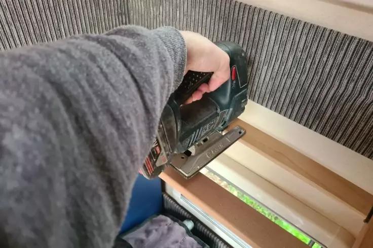 Arm mit grauem &Auml;rmel h&auml;lt eine Stichs&auml;ge &uuml;ber Holzlatten nahe einem Fenster