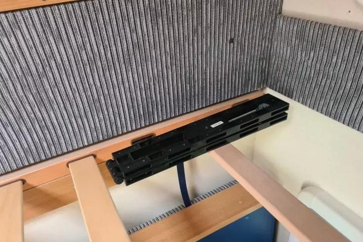 Helle Holzlatten eines Bettgestells, ein schwarzes rechteckiges Bauteil mit blauem Kabel an den Latten, graue vertikal gerippte Wandverkleidung im Hintergrund