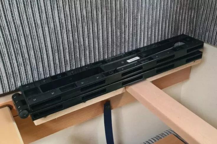 Schwarzer, l&auml;nglicher Tr&auml;ger mit rechteckigen &Ouml;ffnungen liegt auf hellen Holzlatten eines Rahmens; dahinter vertikal gestreifte Wandverkleidung; blauer Gurt h&auml;ngt darunter