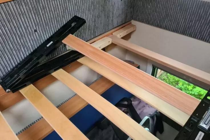 Holzlatten eines Lattenrosts in einem schwarzen Metallrahmen, geriffelte graue Wand im Hintergrund, Taschen unter dem Rahmen