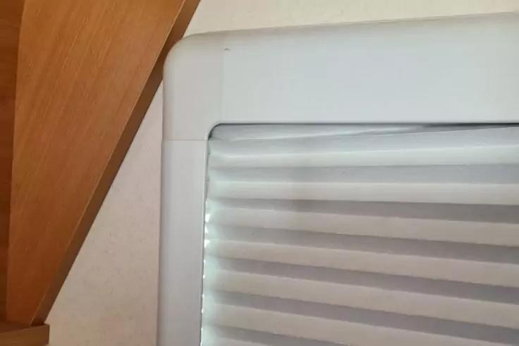 Weißer Fensterrahmen mit beige-weiß gestreiftem Faltstore; links diagonale Holzleiste; weiße Wand