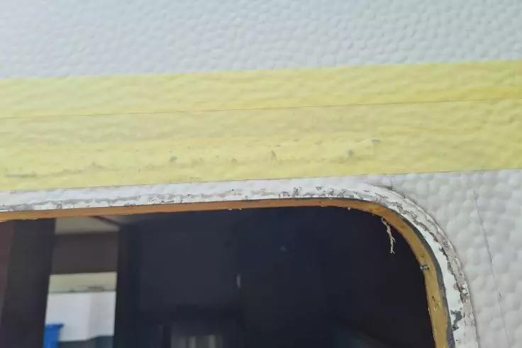 Abgerundetes dunkles Fenster in weißer, strukturierter Fläche mit gelbem horizontalem Streifen oben; dunkle Innenansicht im Glas und kurzer, ausgerissener Faden am Rahmen