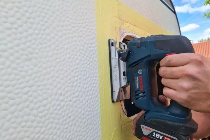 Hand hält eine blaue elektrische Stichsäge an eine gelbe Platte an einer Hauswand, Sägeblatt sichtbar; links weiße strukturierte Wand, oben blauer Himmel