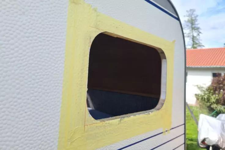 Rechteckige Fensteröffnung in der Seitenwand eines weißen Wohnwagens, Kanten mit gelbem Klebeband, innen dunkel mit sichtbarem blauem Polster; im Hintergrund Haus, Baum und blauer Himmel