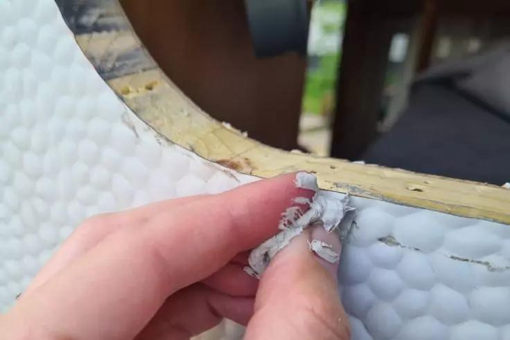 Hand zieht kleine Streifen weißen Schaumstoffs von einer runden, weiß strukturierten Oberfläche mit gelbem Rand; unscharfer Holzhintergrund