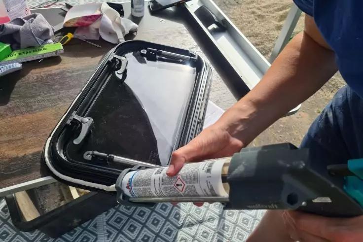 Hände halten eine Kartuschenpistole und tragen Dichtmasse auf einen schwarzen Kunststoffrahmen mit transparenter Abdeckung auf; im Hintergrund Werkzeug und ein gemustertes Tuch