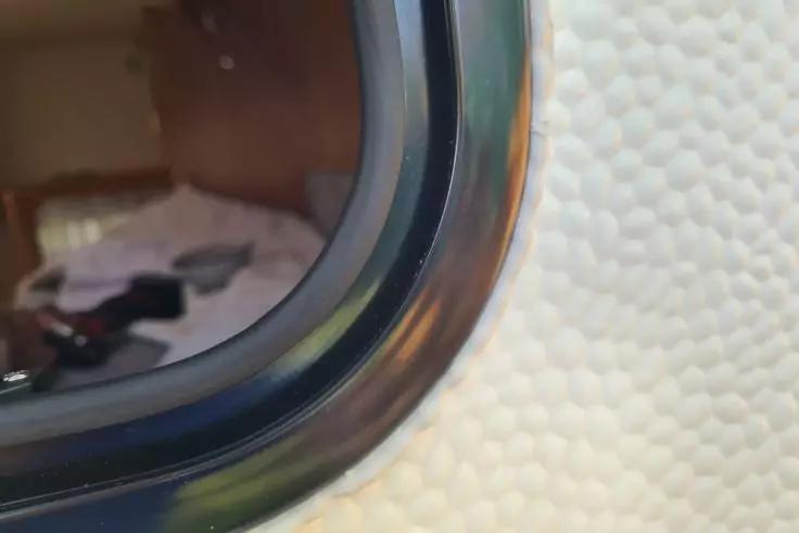 Nahaufnahme eines ovalen Fensters mit schwarzem Rahmen in einer weiß genarbten Wand; im Glas spiegelt sich ein Innenraum mit Bett und Gegenständen