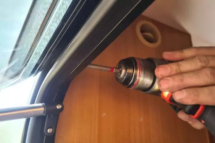 Hand hält Akkuschrauber, Spitze an Schraube im schwarzen Metallrahmen neben Holzverkleidung