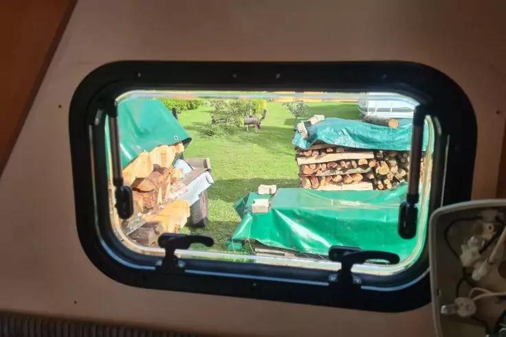 Blick durch ein rechteckiges Fenster auf einen Garten mit gestapeltem Brennholz, teils mit grünen Planen bedeckt, und Rasen