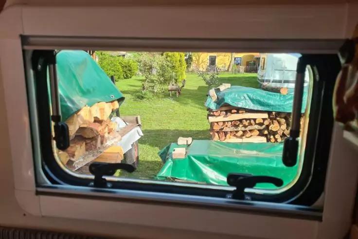 Blick durch ein rechteckiges Fenster mit schwarzem Rahmen und Griffen auf einen Garten mit gestapeltem Feuerholz unter grünen Planen, Rasen, Bäumen und einem gelben Haus im Hintergrund