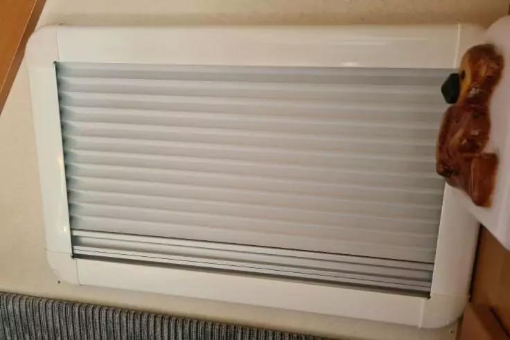 Geschlossenes weißes Rollladenfenster in weißem Rahmen an heller Wand, rechts ein brauner Holzbär an der Rahmenkante, unten graues geripptes Polster