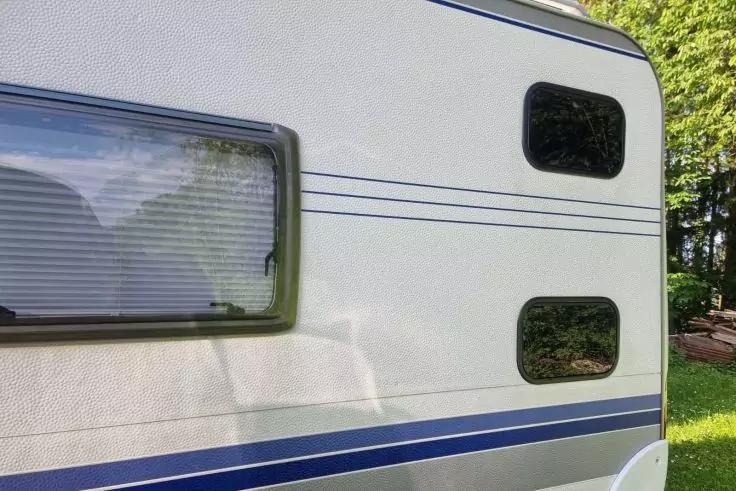 Seitenansicht einer hellen Fahrzeugwand mit drei rechteckigen Fenstern, links geschlossene Jalousie, blaue Streifen, Gras und Bäume im Hintergrund