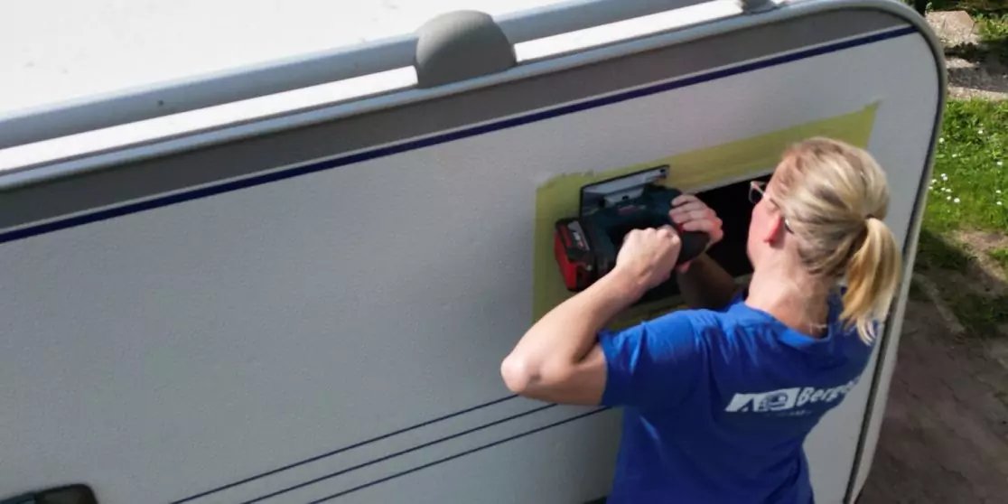 Person mit Pferdeschwanz und blauem T‑Shirt von hinten, h&auml;lt einen Akkubohrer an eine rechteckige &Ouml;ffnung in der Seitenwand eines wei&szlig;en Wohnwagens