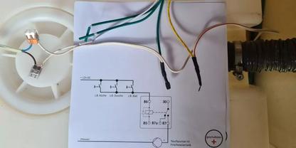 Blatt Papier mit gedrucktem Schaltplan, darauf mehrere isolierte Dr&auml;hte in Gr&uuml;n, Gr&uuml;n-Gelb, Braun, Blau, Schwarz und Wei&szlig;; links eine runde wei&szlig;e Lampenfassung, rechts ein schwarzer Wellschlauch, gelblicher Hintergrund