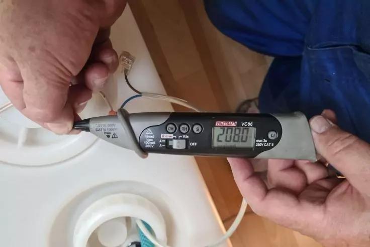 Zwei H&auml;nde halten ein grau-schwarzes handliches Messger&auml;t mit digitalem Display, auf dem "208.0" angezeigt wird; an dem Ger&auml;t sind zwei Messleitungen mit freiliegender Kupferlitze angeschlossen; im Hintergrund ein wei&szlig;es rundes Kunststoffteil und ein