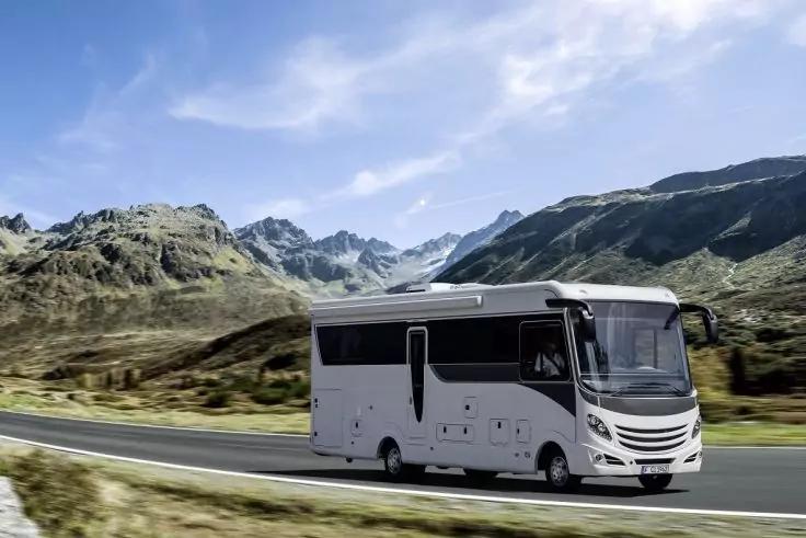 Wei&szlig;es Wohnmobil auf einer asphaltierten Stra&szlig;e vor Bergen und gr&uuml;nen H&uuml;geln unter teilweise bew&ouml;lktem Himmel