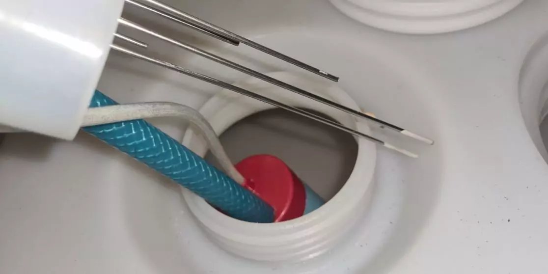 Wei&szlig;e Kunststofffl&auml;che mit zwei runden &Ouml;ffnungen, roter runder Gegenstand in einer &Ouml;ffnung, blaues geflochtenes Rohr f&uuml;hrt hinein, drei d&uuml;nne Metallst&auml;be liegen dar&uuml;ber