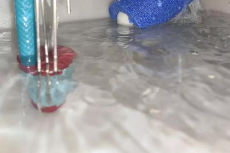 Wasser auf gl&auml;nzender Metallfl&auml;che, drei senkrechte Metallst&auml;be, t&uuml;rkisfarbene Schlauchleitung mit roter Halterung links, blauer Schwamm mit wei&szlig;er Seite oben rechts