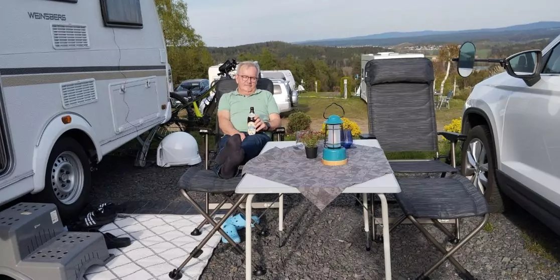 Mann in gr&uuml;nem T-Shirt sitzt an einem Klapptisch zwischen Wohnwagen und Auto; zwei Campingst&uuml;hle, Tisch mit Tischdecke, Thermoskanne und Becher; Landschaft im Hintergrund