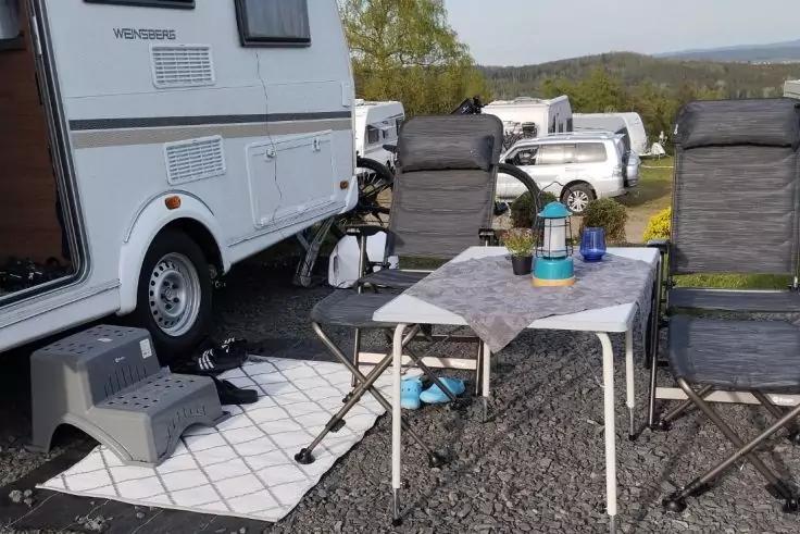 Wohnmobil neben zwei Campingst&uuml;hlen, Campingtisch mit Tischdecke, Thermoskanne und Bechern, Fu&szlig;matte auf Kies, weitere Wohnwagen im Hintergrund