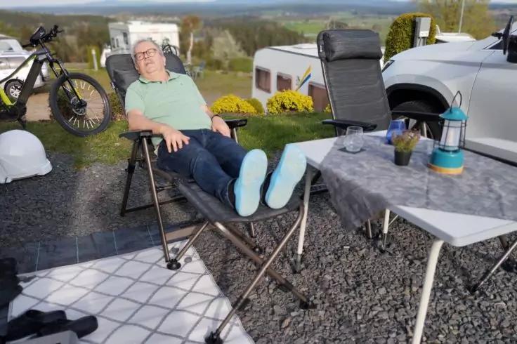 Eine Person sitzt in einem Liegestuhl mit ausgestreckten Beinen auf einer Fu&szlig;st&uuml;tze vor mehreren Wohnwagen; neben ihr ein Tisch mit Laterne und Bechern, links ein Fahrrad, rechts ein Auto, Kiesuntergrund und H&uuml;gel im Hintergrund