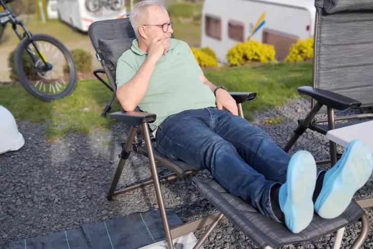 Person mit Brille liegt in einem Camping-Liegestuhl, tr&auml;gt hellgr&uuml;nes T-Shirt und Jeans; Wohnwagen, Fahrradteil, Kiesboden und Gras mit gelben Blumen im Hintergrund