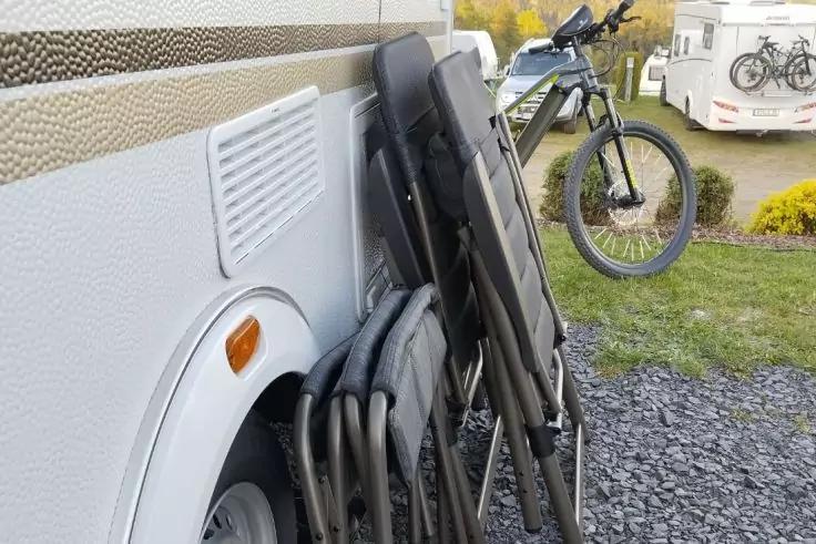 Mehrere zusammengeklappte St&uuml;hle lehnen an der Au&szlig;enseite eines Wohnwagens neben dem Rad; im Hintergrund ein Fahrrad und ein weiterer Wohnwagen