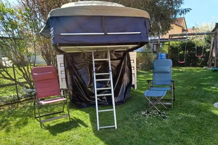 Erh&ouml;hte rechteckige Stoffkonstruktion auf Gestell mit dunklem herabh&auml;ngendem Stoff und einer Leiter davor; links ein roter Klappstuhl, rechts ein blauer Klappstuhl auf Rasen; im Hintergrund B&auml;ume, H&auml;user und eine Schaukel