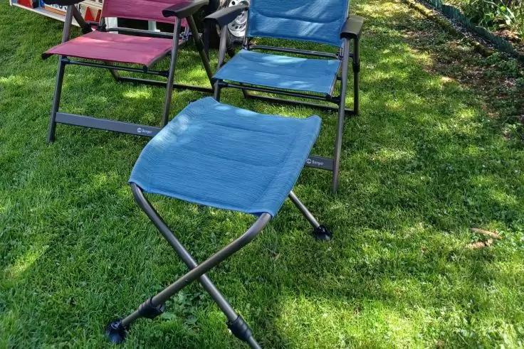 Blauer, zusammenklappbarer Hocker mit Stoffsitz und X-f&ouml;rmigem Metallgestell auf gr&uuml;nem Rasen; dahinter ein blauer und ein roter zusammenklappbarer Stuhl