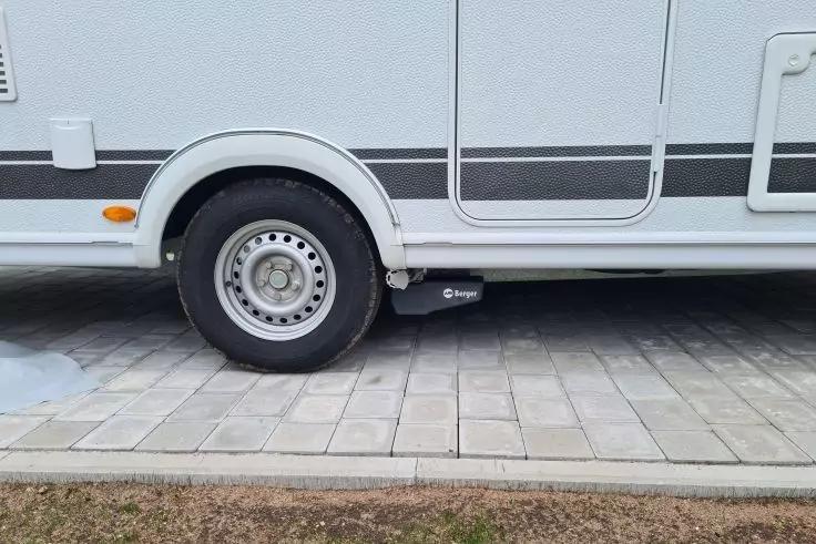 Seitenansicht eines weißen Wohnmobils mit Reifen, Tür, Trittstufe, schwarzem Kasten unter dem Fahrzeug und gepflastertem Parkplatz mit Grasstreifen