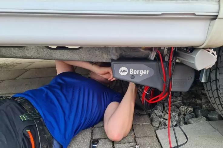 Person unter einem Fahrzeug, trägt blaues T‑Shirt; graues Gerät mit Aufschrift Berger und roten Kabeln, Fahrzeugreifen und Pflastersteine sichtbar