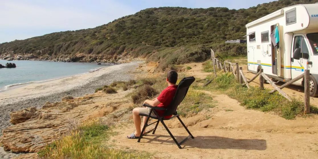 Person in rotem T-Shirt sitzt auf einem Klappstuhl an einem sandig-felsigen Strand und blickt aufs Meer; Wohnmobil steht rechts auf einem Weg