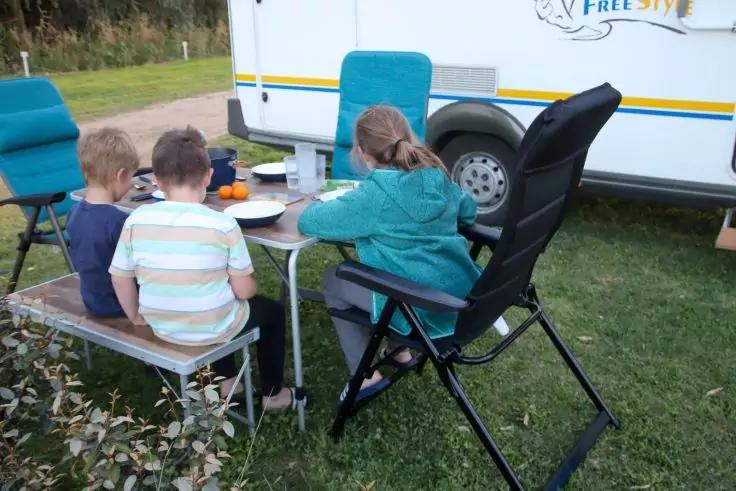 Drei Kinder von hinten an einem Campingtisch auf Gras neben einem wei&szlig;en Wohnwagen; zwei auf einer Holzbank, eines auf einem schwarzen Klappstuhl; Teller und Becher auf dem Tisch