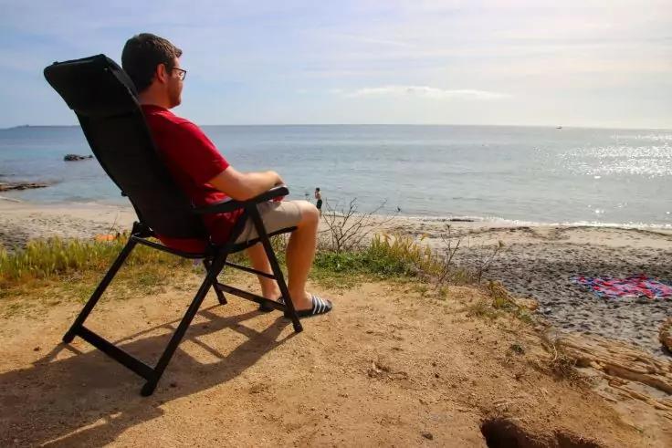 Person sitzt auf Klappstuhl am Strand, Richtung Meer, tr&auml;gt rotes T‑Shirt, kurze Hose und Sonnenbrille