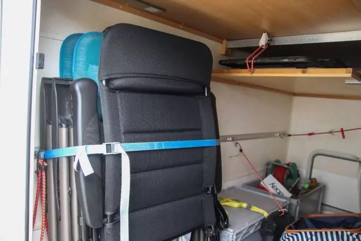 Gefaltete schwarze Sitze, mit blauem Spanngurt befestigt; daneben Regalbretter, Plastikboxen, Werkzeugkiste und gestreifte Tasche