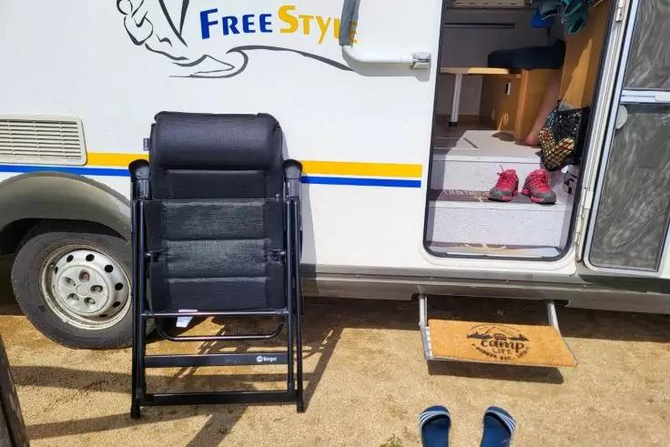 Seitenansicht eines Wohnmobils mit offenem Eingang und Schriftzug "FreeStyle", schwarzem Klappstuhl vor der T&uuml;r, kleinem Holztritt mit Logo, zwei blaue Hausschuhe drau&szlig;en und zwei pinke Hausschuhe im Eingang