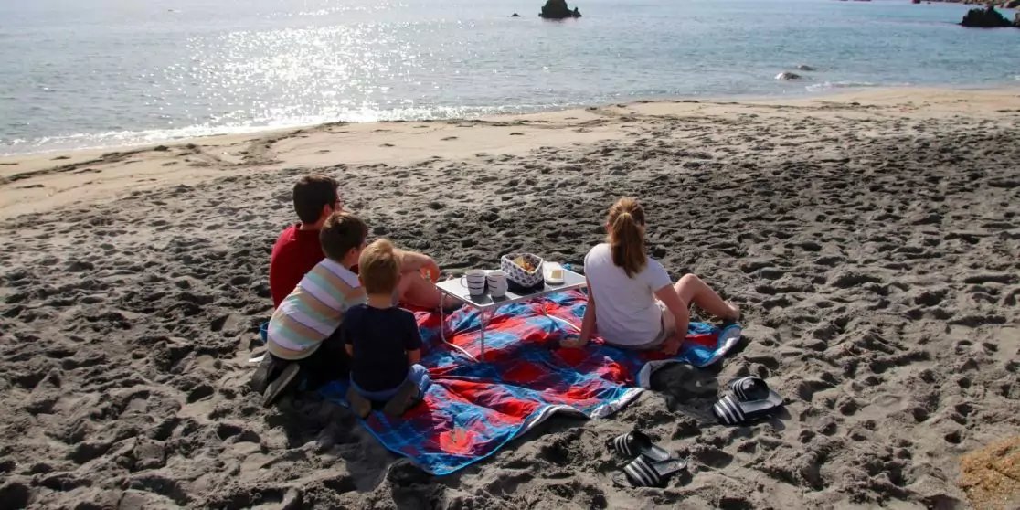 Vier Personen sitzen auf einer rot-blauen karierten Decke auf einem kiesigen Strand, mit Blick zum Meer. Sonnenreflexe auf dem Wasser, Felsen im Meer, Schuhe und Tasche neben der Decke