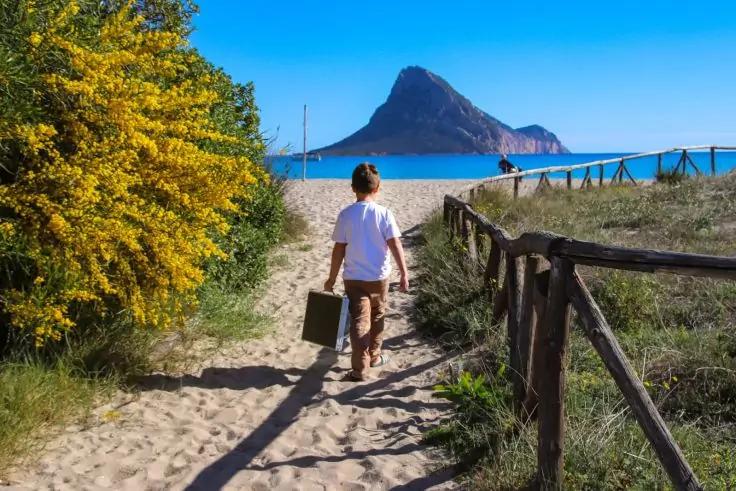 Person von hinten in wei&szlig;em T‑Shirt und kurzen Hosen tr&auml;gt einen Koffer auf einem sandigen Weg zum Strand; links bl&uuml;hender gelber Busch, rechts Holzzaun, im Hintergrund ein markanter Felsen im Meer, blauer Himmel