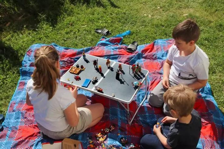 Drei Kinder sitzen auf einer karierten Picknickdecke auf dem Rasen; ein kleiner Klapptisch mit Spielzeugfiguren steht vor ihnen; weitere Spielzeuge liegen auf der Decke