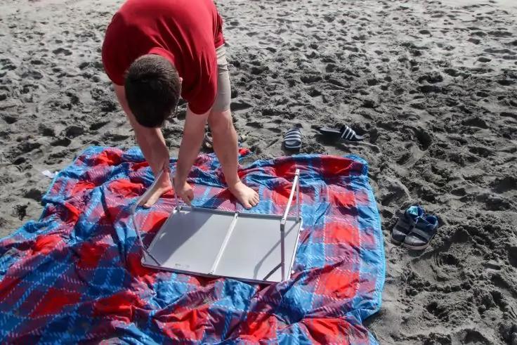 Auf einem blau-roten Strandtuch auf dunklem Sand beugt sich eine Person in rotem T-Shirt &uuml;ber einen kleinen silbernen Klapptisch; rechts liegen zwei Sandalen